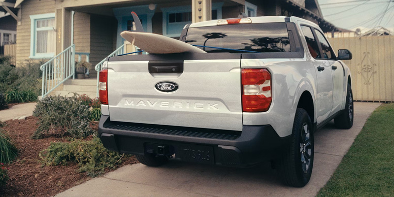 A white Ford Maverick with a surfboard secured on its back end, parked outdoors.